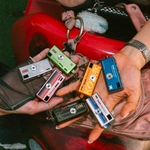 Several miniature film camera keychains in different colors are held in two hands over a red motorcycle tank and a pink bag. The keychains have vintage retro designs with text such as 'KODAK', 'FIALE', 'Spiral', and 'Mini Film Camera'.