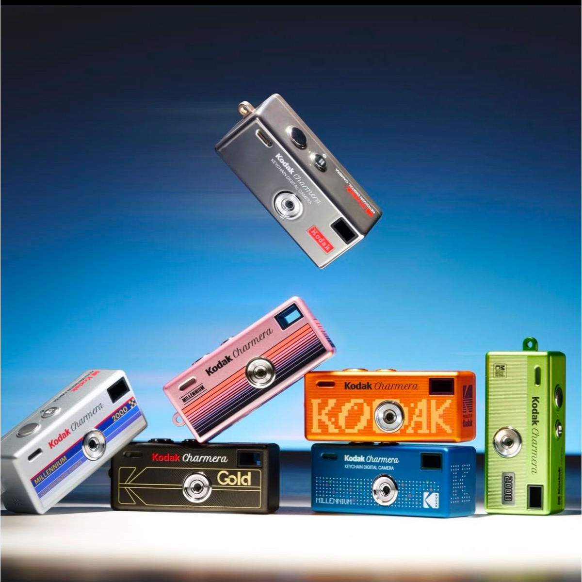 Six Kodak Charmars instant cameras in assorted colors, including silver, pink, orange, black, blue, and green, arranged dynamically against a blue gradient background.